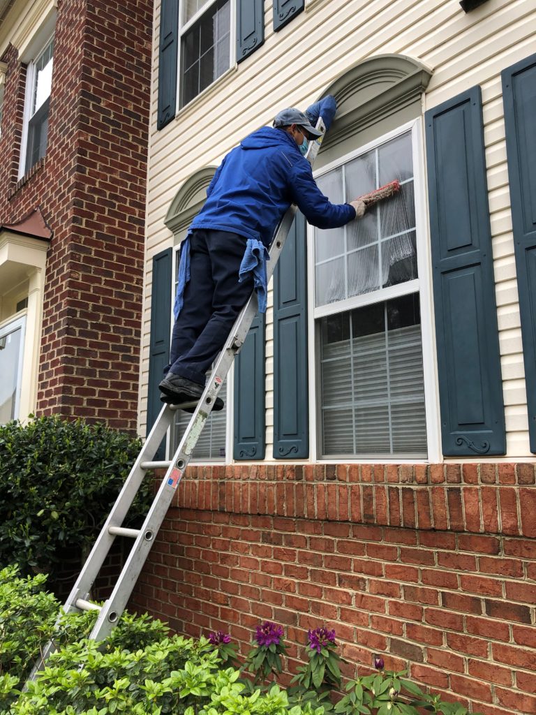 Technician on a ladder, cleaning the top of a double-hung window from the outside.